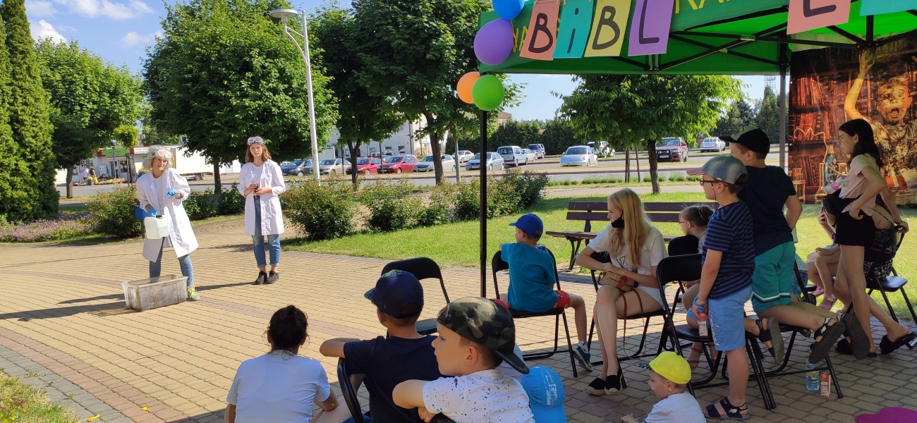 Zdjęcie kolorowe, uczestnicy zabawy pod hasłem Wiosna z doświadczeniami, w tle szalony profesorek z pomocnikiem robiący dymiący eksperyment, plac przed Biblioteką